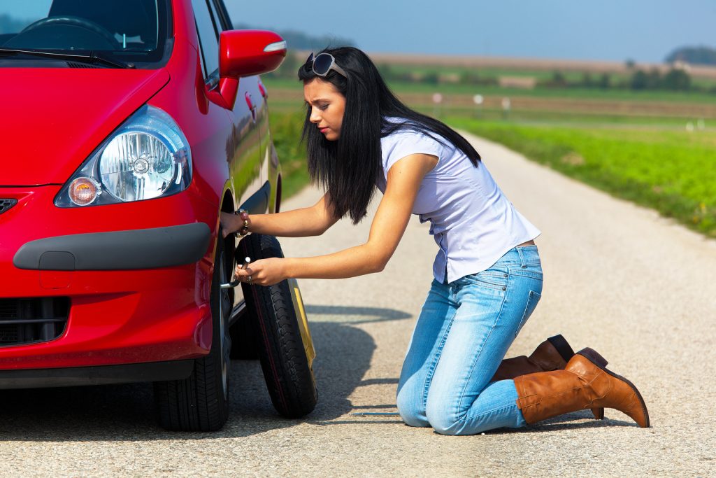 Young woman crouched down and changing a tire on her car. Horizontally framed shot.