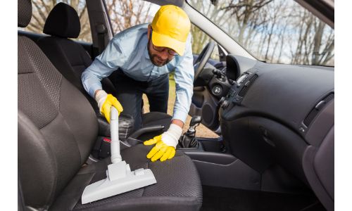 a man with a yellow hat and gloves vacuuming a vehicle's seating upholstery